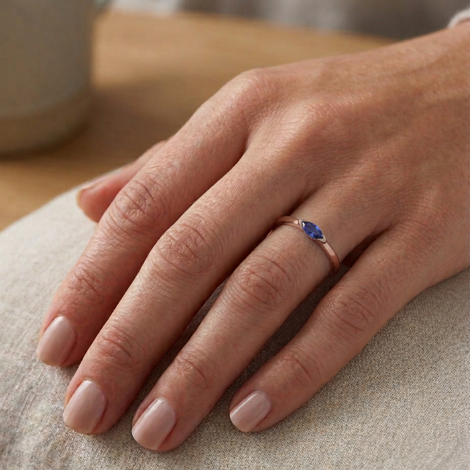 Hand wearing a slim rose gold ring with a blue sapphire on a neutral background